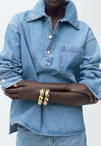 Osoba w jasnoniebieskiej koszuli dżinsowej i dżinsach, z założonymi rękami, mająca na lewym nadgarstku dwa masywne złote bransoletki.