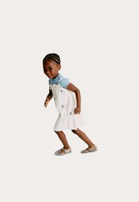 Hvid broderet kjole med en flæset kant, båret over en lyseblå skjorte. Barnet har klare sandaler på og poserer i en legesyg stilling.