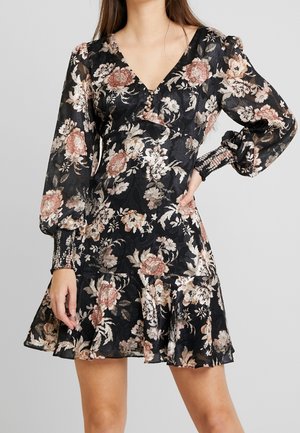 Femme portant une robe noire à fleurs avec des manches longues bouffantes, un col en V, des boutons, des poignets froncés et un ourlet évasé à volants.