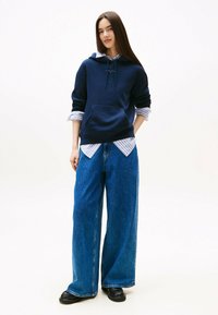 Felpa navy con tasca frontale e cordini, indossata sopra una camicia a righe blu chiaro; abbinata a jeans blu a gamba larga e stivali neri.