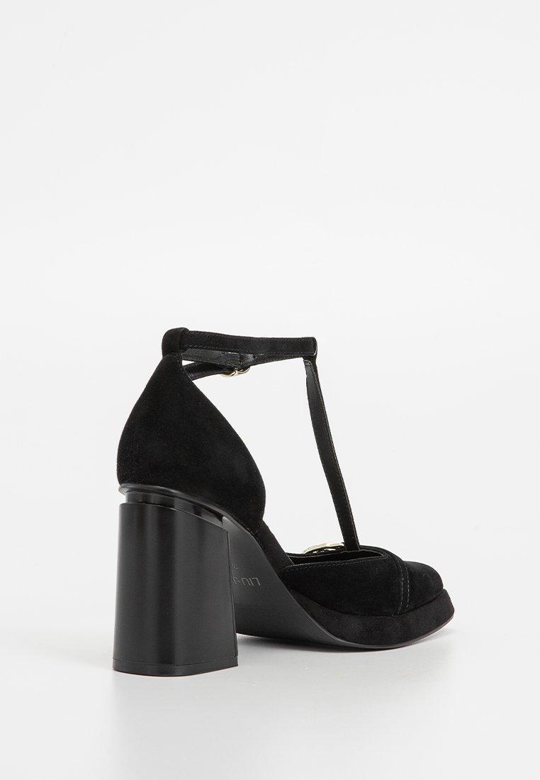 Zapato de tacón alto de ante negro con tira en T, tacón grueso y punta redonda, visto desde la parte posterior y lateral sobre un fondo blanco liso.
