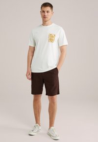 Camiseta blanca de algodón con un gráfico amarillo en el lado izquierdo del pecho, combinada con pantalones cortos marrones y zapatillas blancas, de pie contra un fondo neutro.