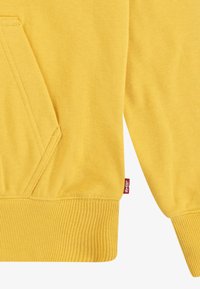 Gul genser med kengurulomme, ribbede mansjetter og hem, myk tekstur, med en liten rød Levi's-lapp på siden.