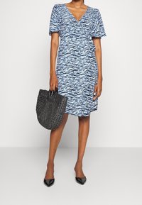 Blå mönstrad omlottklänning med korta puffärmar. Kvinna håller en vävd svart handväska och har på sig spetsiga svarta klackar. Slätt, lättviktsmaterial.