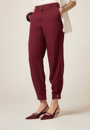 Femme portant un pantalon fuselé bordeaux taille haute avec des poignets à boutons et des escarpins à bride arrière assortis à talons pointus, les mains dans les poches.