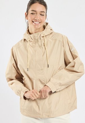 Femme souriante aux cheveux foncés portant une veste à capuche beige, les mains jointes devant, regardant légèrement vers le bas et sur le côté.