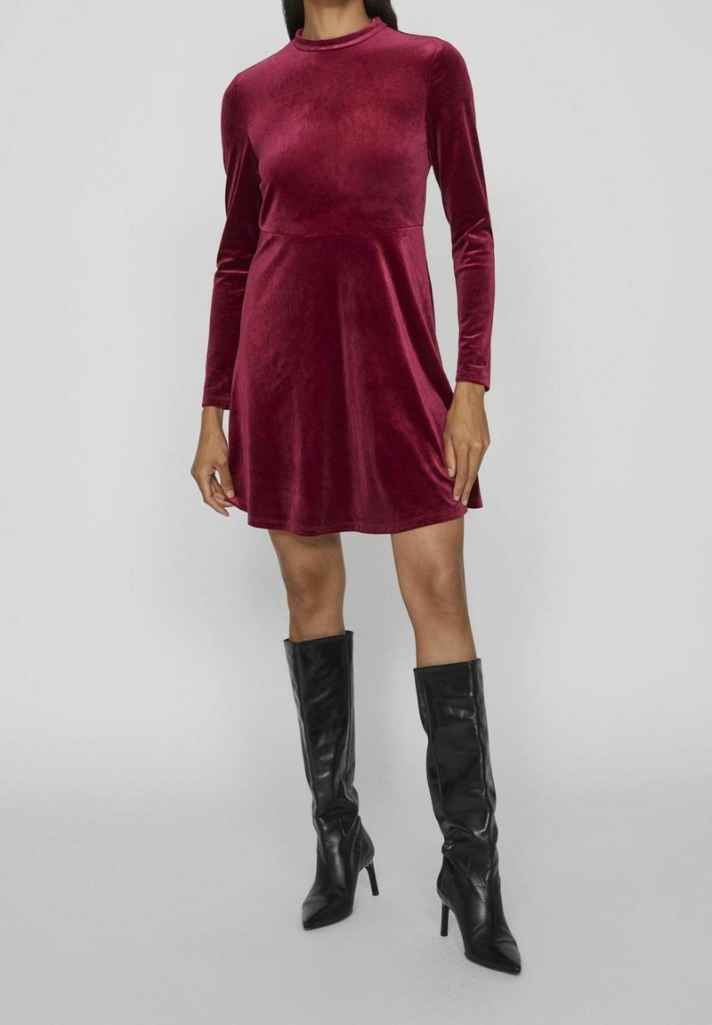 Burgundy sammet långärmad klänning med figurnära överdel och utfälld kjol, paired med knähöga svarta läderstövlar med klack.