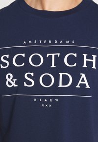 Granatowa bawełniana koszulka z dużym białym napisem "SCOTCH & SODA" oraz mniejszymi napisami "AMSTERDAMS" i "BLAUW." Krótkie rękawy.
