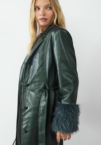 Ciemnozielony skórzany płaszcz z pasek w talii, z futrzanymi akcentami przy mankietach. Gładka tekstura i klasyczny podwójny guzik.