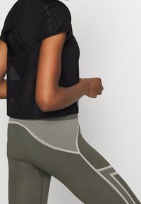 Mustat lyhythihaiset crop-topit, joissa on läpinäkyvä selkä. Oliivinvihreissä leggingseissä on verkkopaneelivuorauksia ja kaarevia kuvioita vaaleammissa sävyissä.