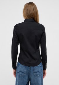 Black long-sleeve shirt with a collar, featuring a smooth texture and structured back design. Worn with blue jeans with detailed stitching.