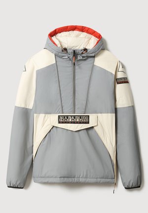 Personne portant une veste à capuche grise et beige avec poche avant, un pantalon en polaire noir et des bottes noires sur un fond uni.
