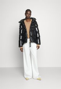 Czarna, błyszcząca puchowa kurtka z kapturem, zapinana na zamek, z elastycznymi detalami, noszona na beżowej bluzce i zestawiona z szerokimi białymi spodniami.