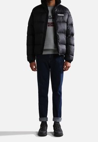 Giacca a vento nera con collo alto, chiusura con zip e dettaglio del logo, indossata sopra una felpa grigia, abbinata a jeans blu scuro e scarpe nere.