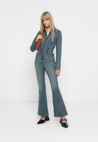Combinaison en denim bleu délavé avec un corsage ajusté, des jambes évasées et un col contrastant. Portée avec des chaussures à talons carrés noires et un sac à main marron.