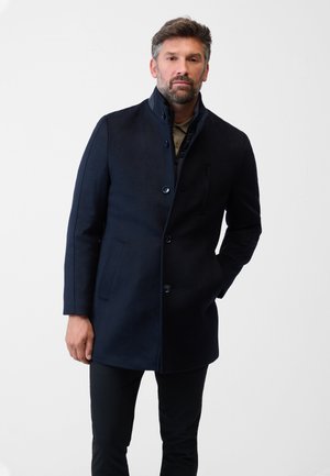 Navy coat made of wool blend, featuring a high collar, button closure, side pockets, and a streamlined fit over dark pants.