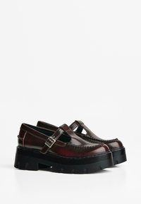 Braune Lederschuhe mit einem T-Riemen, dicker schwarzer Plattformsohle und dekorativen Nähten entlang der Kante. Glatte Oberfläche, minimales Zubehör.