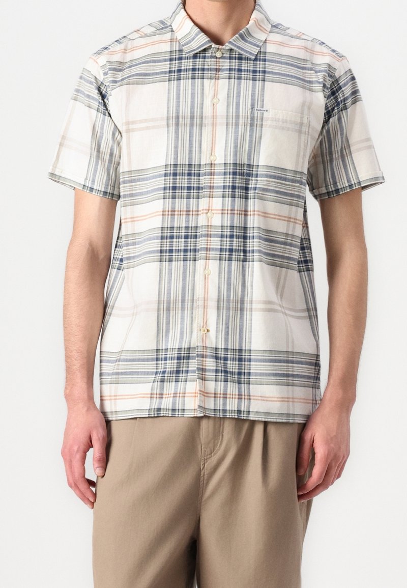 Homme portant une chemise à manches courtes boutonnée avec un motif à carreaux blanc, bleu et orange et un pantalon beige sur fond blanc.