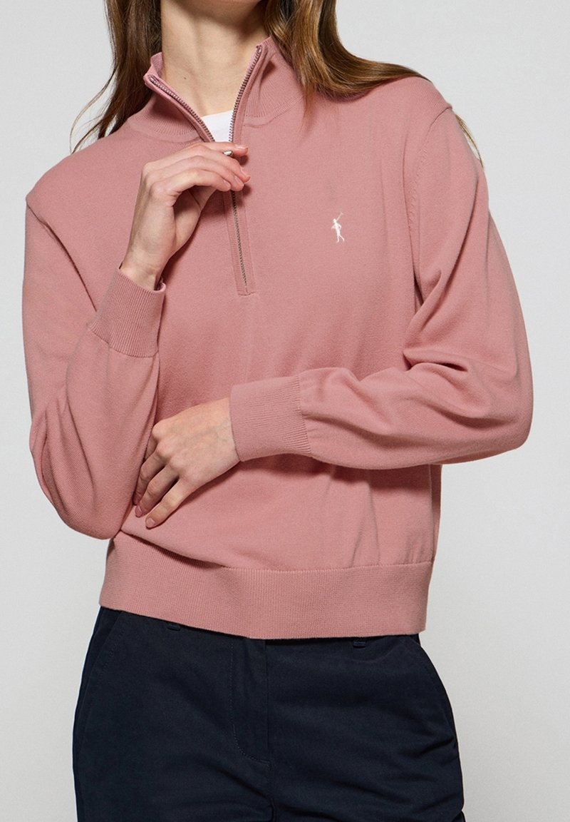 Femme portant un pull rose poudré à fermeture éclair partielle avec poignets et ourlet côtelés, associé à un pantalon bleu foncé, ajustant la fermeture éclair.