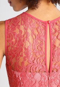 Röd spetsklänning med blommönster, genomskinlig rygg och knäppning vid halsen. Slät tyg nedanför spetsdetaljerna.