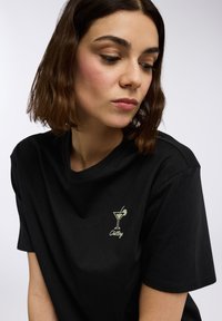 Maglietta nera in cotone con maniche corte e scollatura rotonda. Design ricamato in oro di un bicchiere da cocktail con la scritta "Chilling".