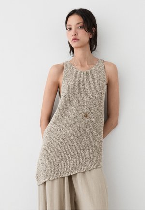 Jeune femme portant un top en maille beige sans manches et un pantalon ample beige clair, debout contre un mur blanc uni.