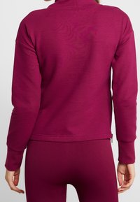 Přiléhavá mikina v hluboké purpurové barvě, s dlouhými rukávy, vysokým límcem a zipem na zadním lemu. Měkká, strukturovaná látka.