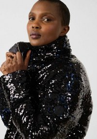 Femme aux cheveux courts portant une veste noire brillante à paillettes, tenant le col près de son visage, regardant avec confiance devant elle.