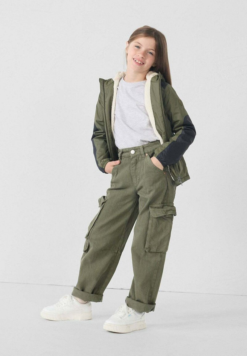 Giacca con cappuccio verde oliva con fodera in pile bianco, maglietta grigia e pantaloni cargo. Scarpe da ginnastica bianche con suole testurizzate. Vestibilità comoda, tasche funzionali.