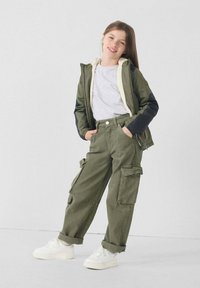 Olivengroene gewatteerde jas met capuchon en witte fleece voering, grijs T-shirt en cargobroek. Witte sneakers met gestructureerde zolen. Losse pasvorm, functionele zakken.