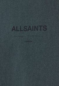 Tamsiai žalia medvilninė marškinėlė su grublėta tekstūra. Dėl marškinėlių dizaino naudojamas juodas ekranu spausdintas tekstas "ALLSAINTS" ir "LONDON". Paprastas, minimalistinis dizainas.