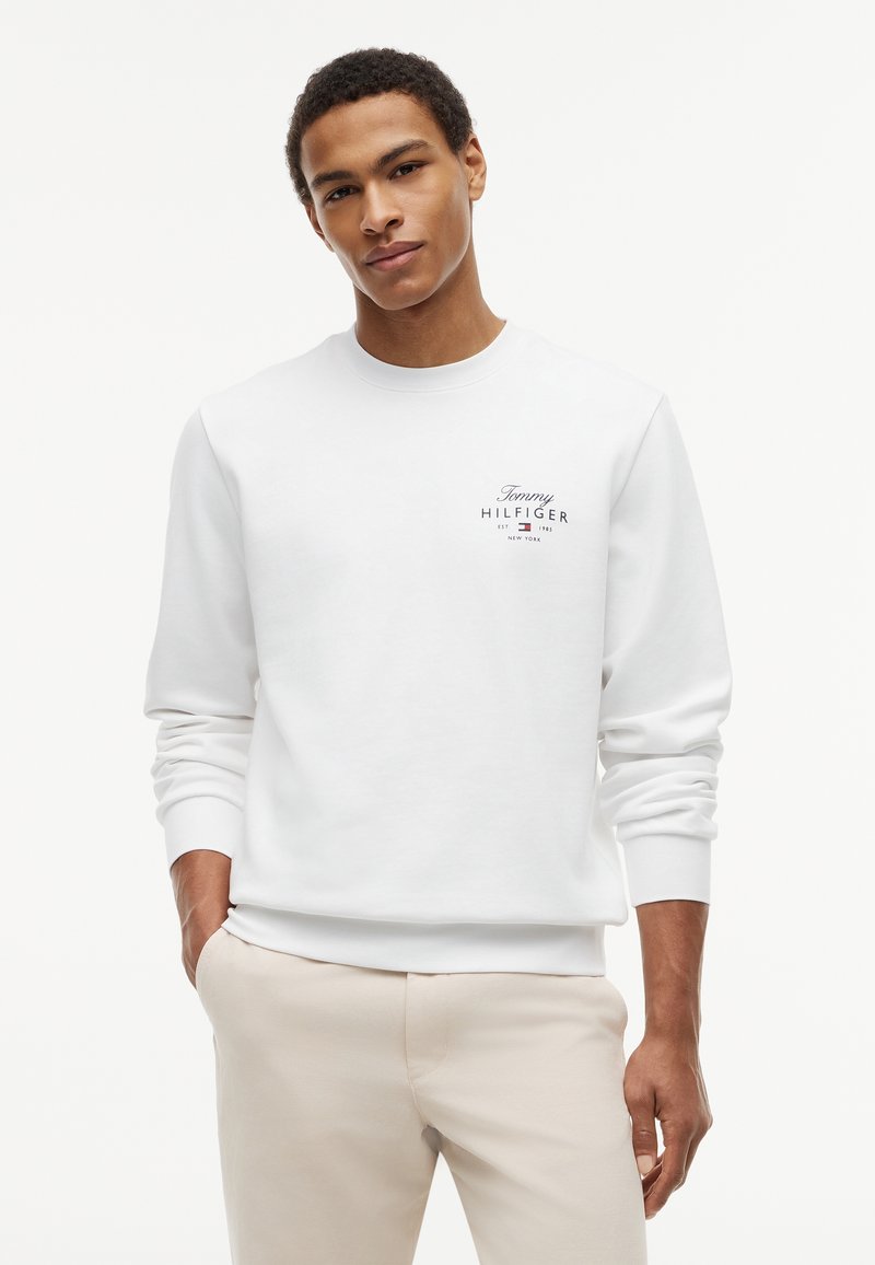 Jeune homme debout, mains dans les poches, portant un sweat-shirt blanc Tommy Hilfiger et un pantalon beige clair sur un fond uni.