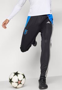 Juodi sportiniai kelnės su mėlynomis šoninėmis juostelėmis, su baltais dryžiais ir logotipu. Dėvimi su balta ilgomis rankovėmis palaidine ir juodais sportbačiais.
