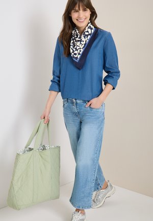 Femme portant un haut bleu, une écharpe à motifs, un jean bleu clair et des baskets argentées, tenant un grand sac fourre-tout matelassé vert clair.