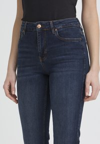 Jean bleu foncé taille haute avec bouton en cuivre et coutures apparentes, porté par une personne en haut noir, visible de la taille jusqu'à mi-cuisse.