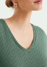 Grön texturerad topp med V-ringning, med ett fiskbensmönster. Guldhoopörhängen synliga, som framhäver detaljer och material.