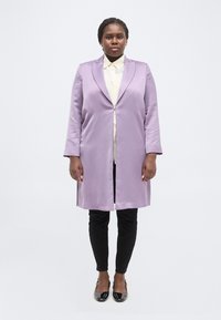 Γυναίκα που στέκεται κοιτώντας μπροστά, φοράει ένα μακρύ μωβ παλτό, λευκό πουκάμισο, μαύρο παντελόνι και μαύρα παπούτσια από γυαλιστερό δέρμα, σε απλό λευκό φόντο.