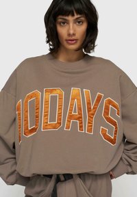 Brązowy, oversize'owy sweatshirt z pomarańczowym, satynowym napisem "100 DNI". Posiada okrągły dekolt i luźny krój z elastycznymi mankietami.
