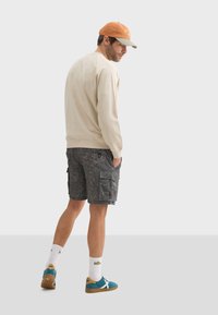 Sweat-shirt beige, short cargo gris avec motif de feuilles, baskets teal avec accents beige, et casquette orange et grise portée à l'envers.