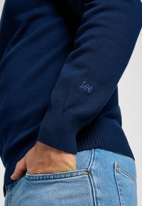 Granatowy sweter robiony na drutach, z prążkowanymi mankietami i dołem, ozdobiony małym, jasnoniebieskim haftowanym logo na rękawie. Casualowy krój.