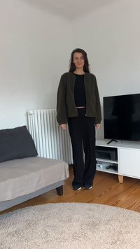 Kvinde står i stuen iført sorte bukser, sort top og mørk jakke nær en grå sofa og et hvidt TV-bord med TV.