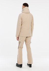 Beige wasserdichte Jacke mit hohem Kragen, verstellbarem Kapuzen und Seitentaschen. Kombiniert mit passenden Hosen mit Reißverschlüssen an den Knöcheln.