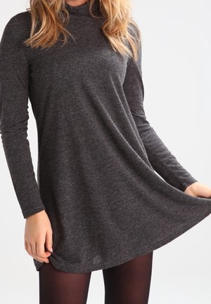 Kvinde iført en langærmet, mørkegrå rullekravekjole, der holder i kjolens kant, mens hun har sorte strømpebukser på, foran en ensfarvet baggrund.