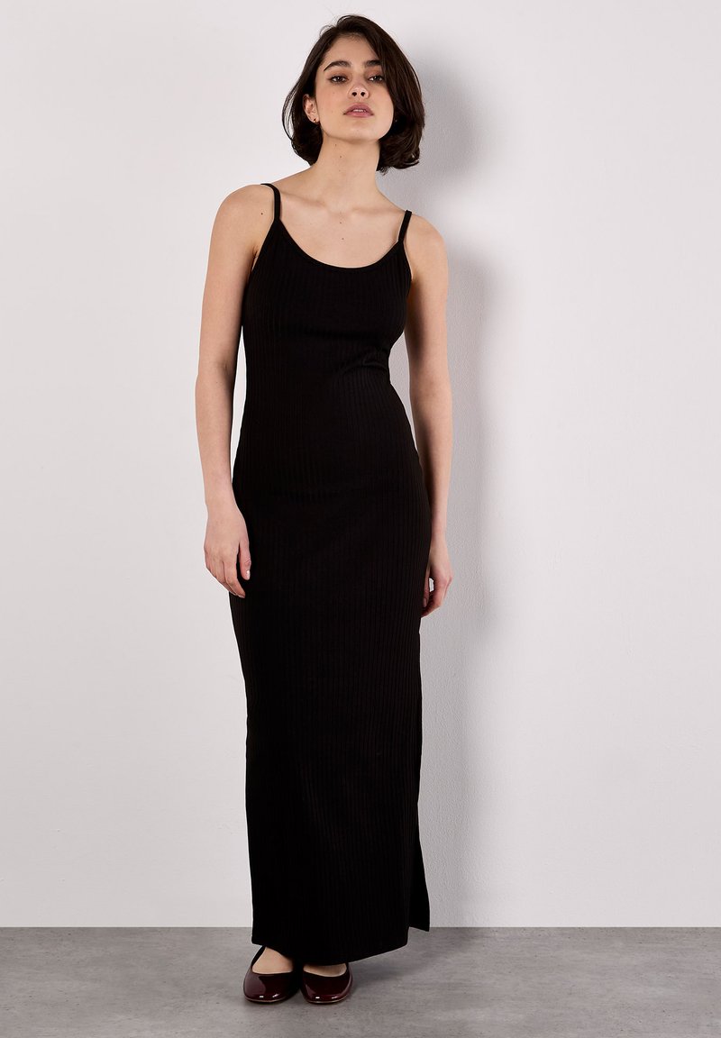 Abito maxi nero a costine con spalline sottili, forma aderente e spacco laterale. Accompagnato da scarpe basse bordeaux su uno sfondo neutro.