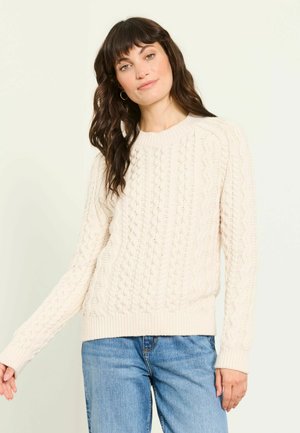 Kvinde med mørkt bølget hår, iført guldringe-øreringe, en cremefarvet kabelstrikket sweater og blå jeans mod en lys baggrund.