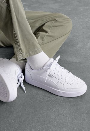 Osoba nosząca białe buty sportowe Puma, białe skarpetki i spodnie w kolorze khaki, siedząca ze skrzyżowanymi nogami na szarej podłodze.