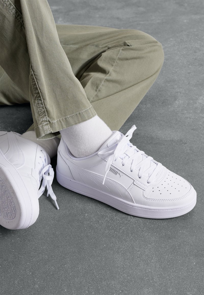 Personne portant des baskets blanches Puma, des chaussettes blanches et un pantalon kaki, assise jambes croisées sur un sol gris.