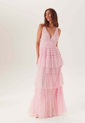 Kvinna som ler, klädd i en ärmlös, rosa maxiklänning med volanger och djup V-ringning, stående mot en enkel ljus bakgrund.