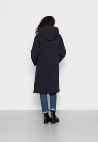 Abrigo largo acolchado negro con capucha, con costuras horizontales, combinado con vaqueros azules y botas negras. Textura lisa, diseño práctico.