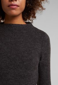 Femme aux cheveux bouclés portant un pull en laine gris foncé à manches longues, visible du cou au haut du torse sur fond gris.